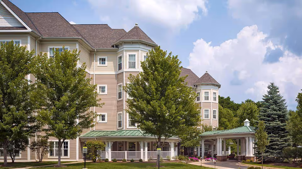 Exterior view of a multi-story senior living facility building with beige siding and a green metal roof over the entrance. The building is surrounded by green trees and landscaping under a partly cloudy sky.