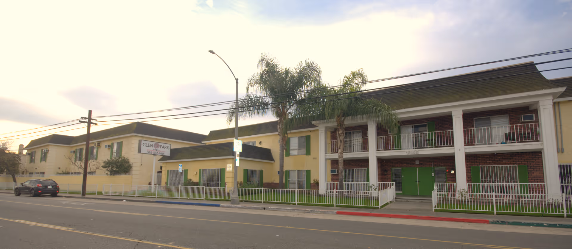 Exterior view of Glen Park at Long Beach, a two-story senior living facility with beige and brick walls, green window shutters, white railings, and palm trees in front. There is a sign with the facility name near the entrance and a car parked on the street.