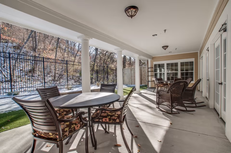 Covered outdoor patio area with multiple seating options including a round table with four chairs and several wicker rocking chairs. The patio is supported by white columns and overlooks a fenced area with trees and a light dusting of snow on the ground.