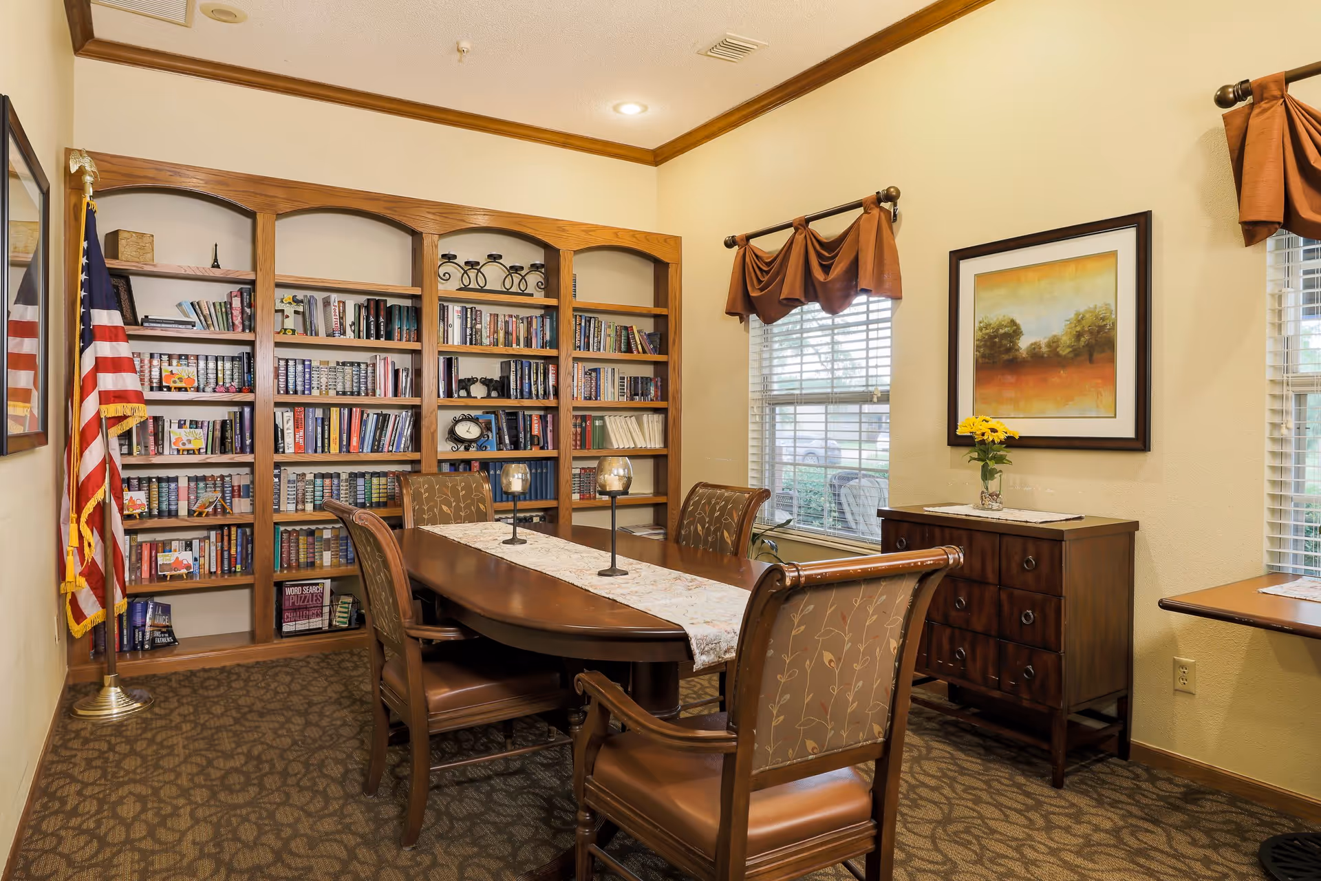 A cozy interior room with a wooden table surrounded by four upholstered chairs. Behind the table is a large wooden bookshelf filled with books and decorative items. An American flag stands in the corner. The room has beige walls, two windows with brown valances, a wooden chest of drawers with a vase of yellow flowers, and a framed landscape painting on the wall.