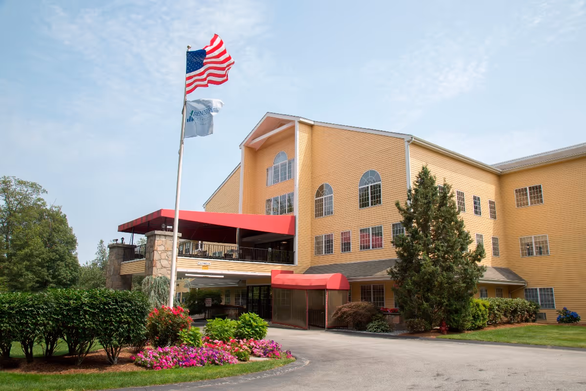 Exterior view of a multi-story yellow building with large windows and a red awning over the entrance. There are two flagpoles with an American flag and another flag, surrounded by green bushes and colorful flowers under a partly cloudy sky.