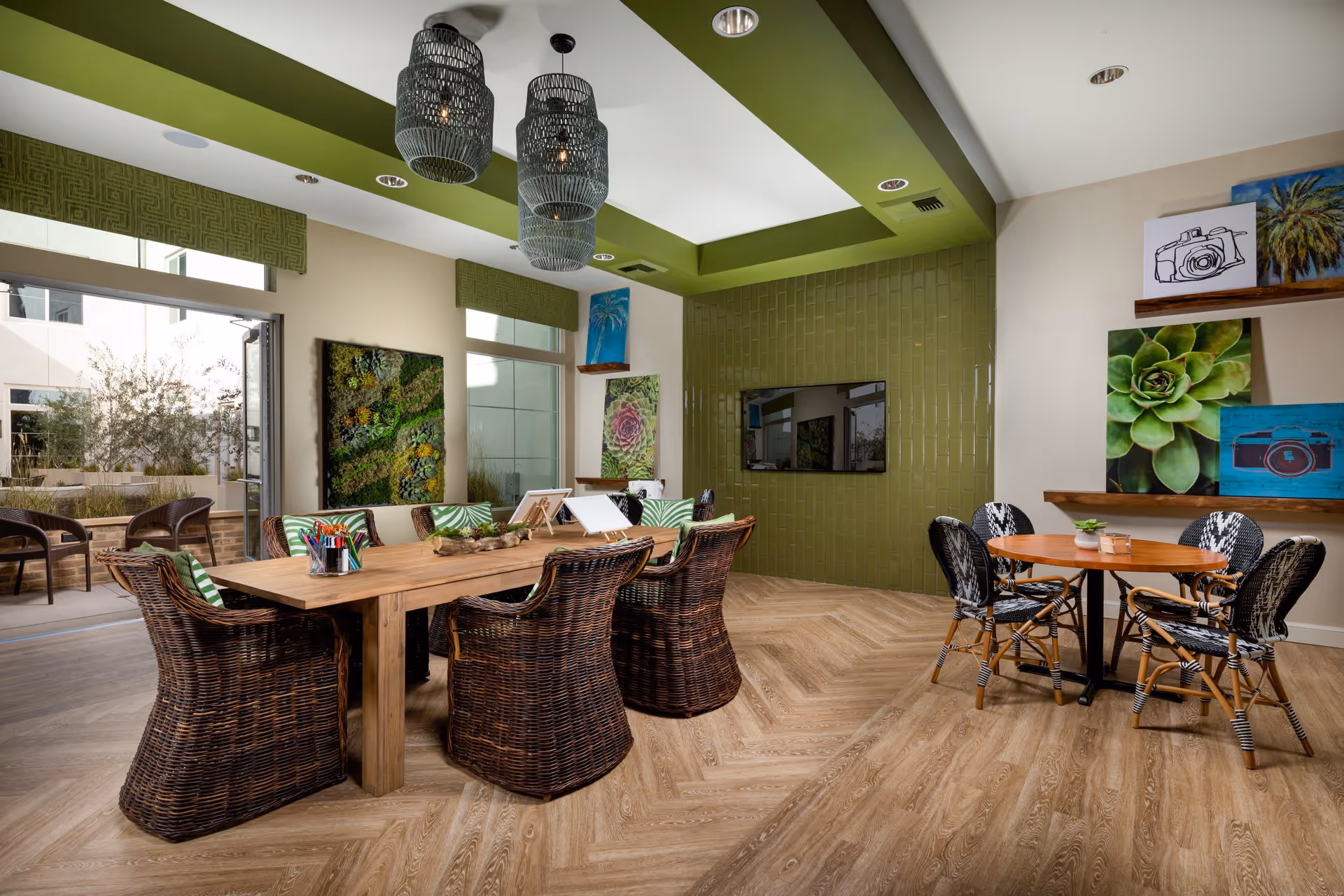 A bright and modern common area with a large wooden table surrounded by wicker chairs with green and white cushions. There is a smaller round table with four black and white patterned chairs. The room features green accent walls, decorative hanging lights, and various colorful artwork including images of succulents and cameras. Large windows and a glass door open to an outdoor patio with additional seating.