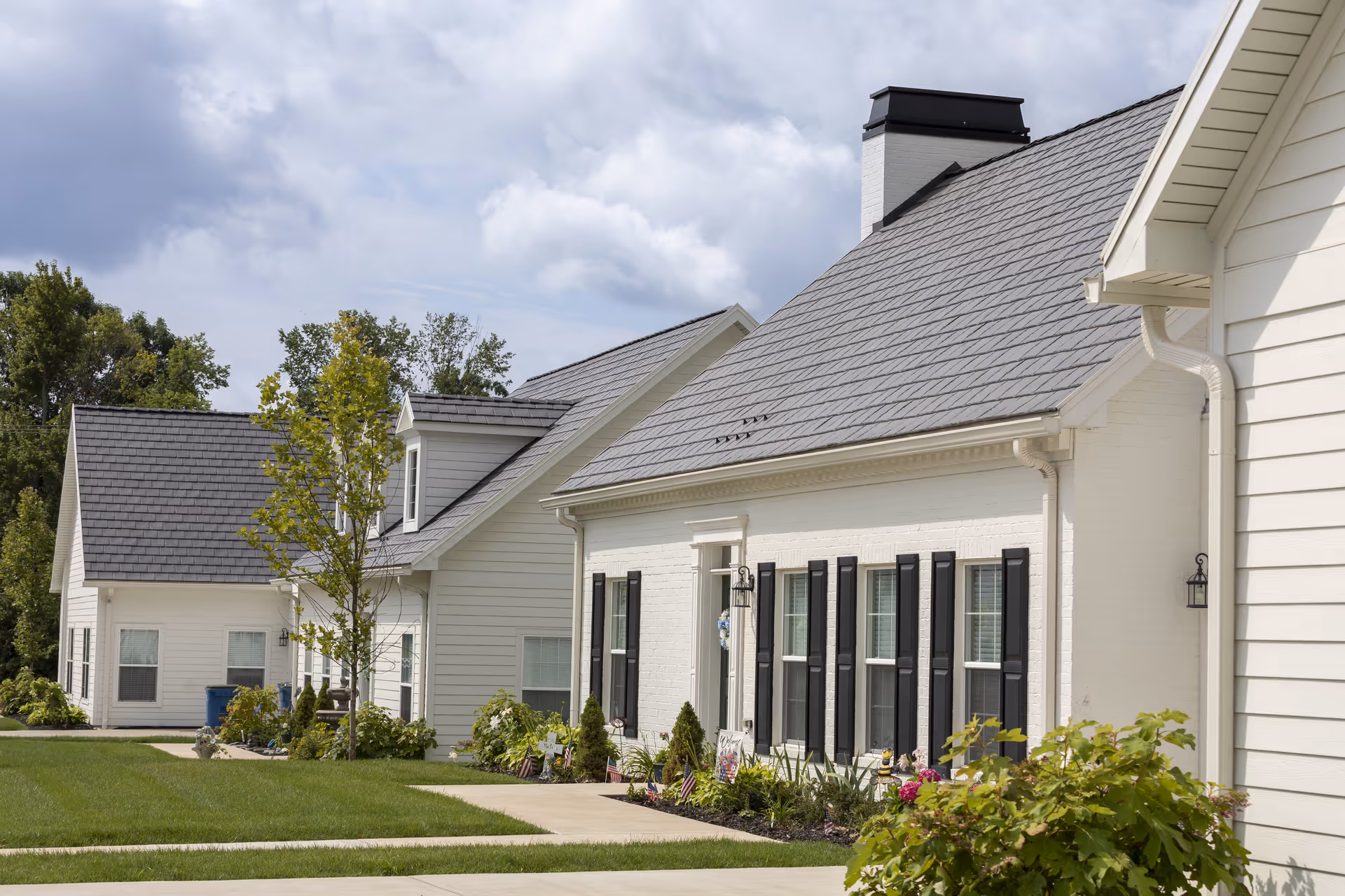 Exterior view of a senior living facility showing white buildings with gray roofs, black window shutters, and well-maintained landscaping including green grass, small trees, and flower beds under a cloudy sky.