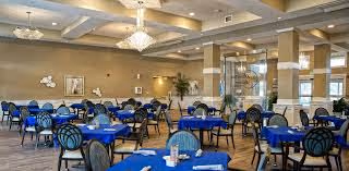 Spacious dining room with multiple round tables covered with blue tablecloths and set with napkins and utensils. The room features beige walls, large windows allowing natural light, decorative wall art, and elegant chandeliers hanging from a coffered ceiling.