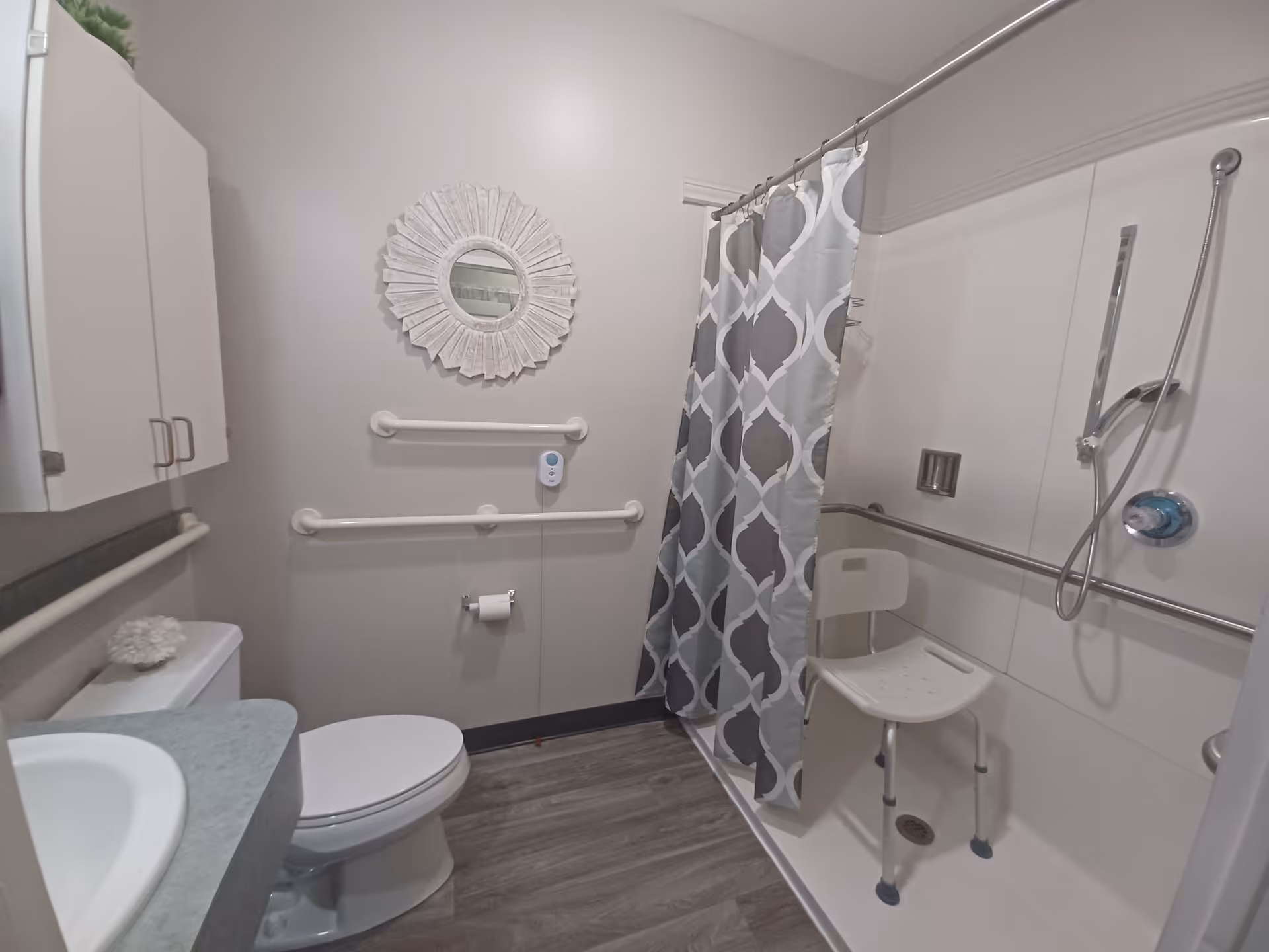 A bathroom with a toilet, sink with countertop, wall-mounted cabinet, and a shower area with a shower chair and a patterned shower curtain. The walls have grab bars for accessibility, and there is a decorative round mirror above the toilet.
