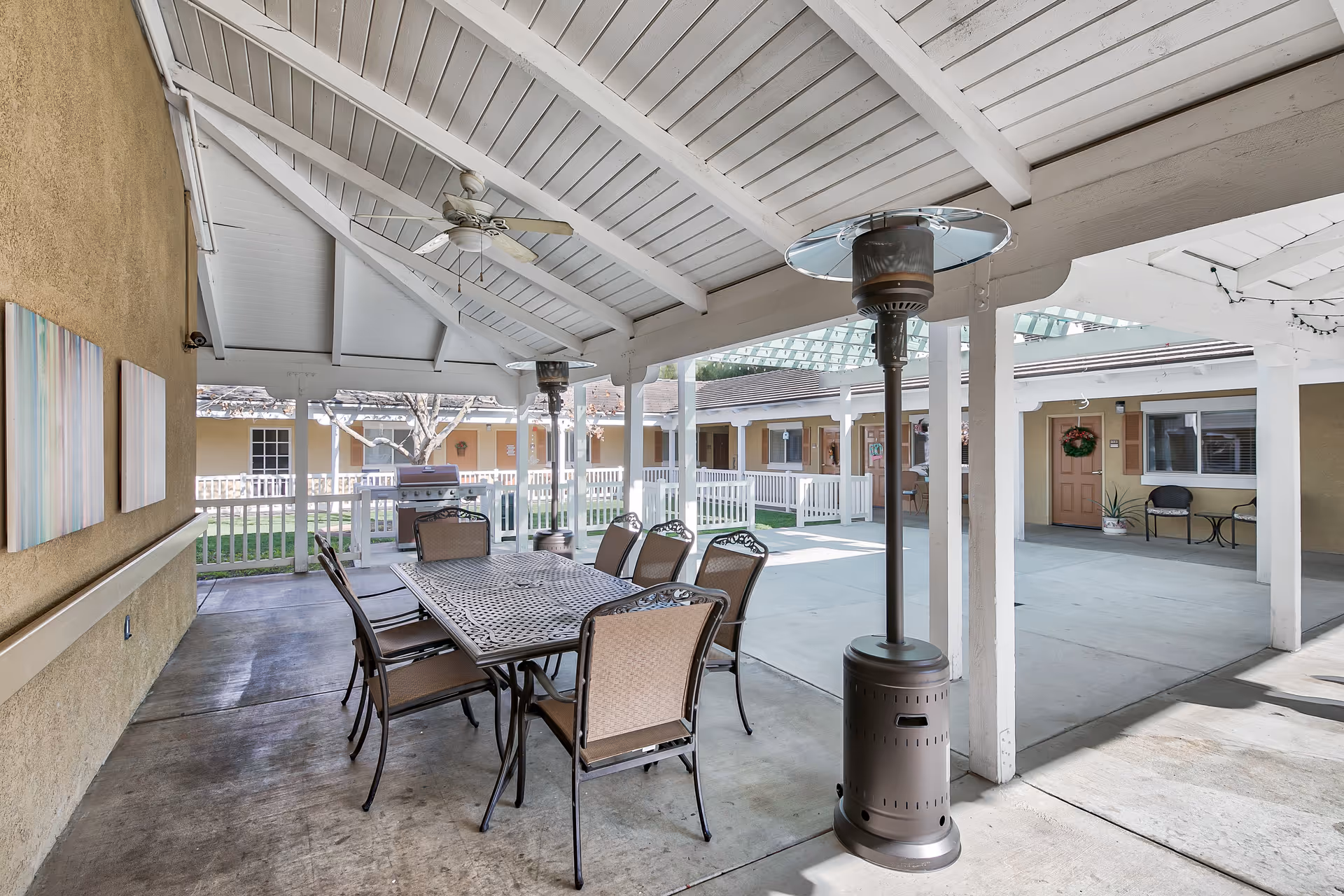 Covered outdoor patio area with a long metal table and eight chairs. Two tall outdoor heaters are positioned near the table. The patio has a white wooden ceiling with a ceiling fan. In the background, there is a fenced grassy courtyard and a building with multiple doors and windows.