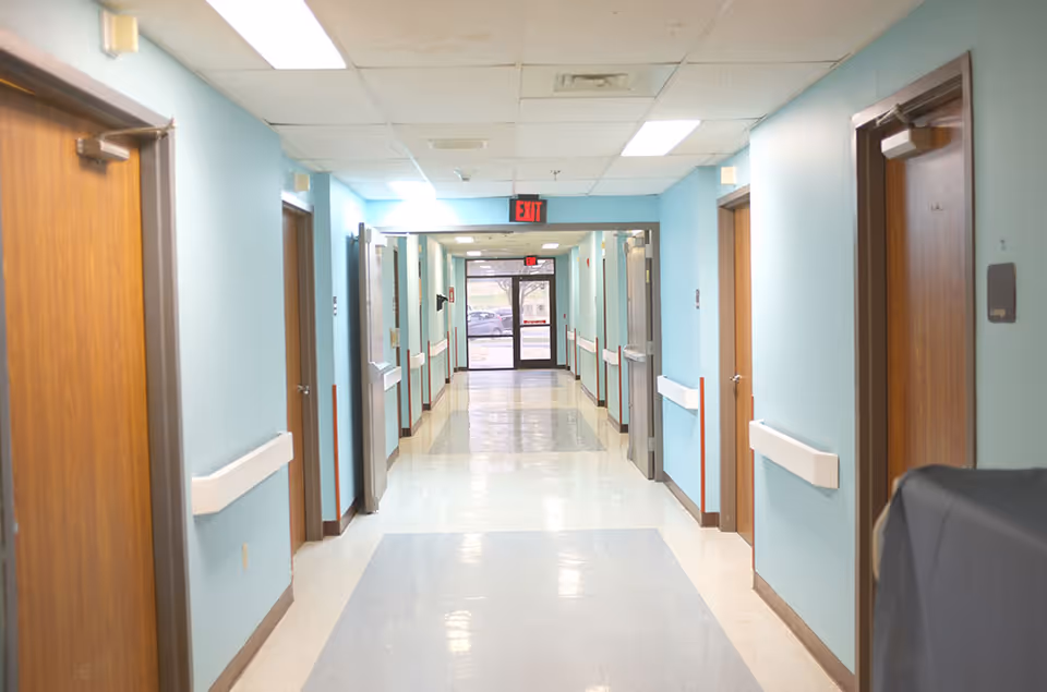 A clean, well-lit hallway in an assisted living facility with light blue walls, wooden doors on both sides, white handrails, and a glass exit door at the far end.
