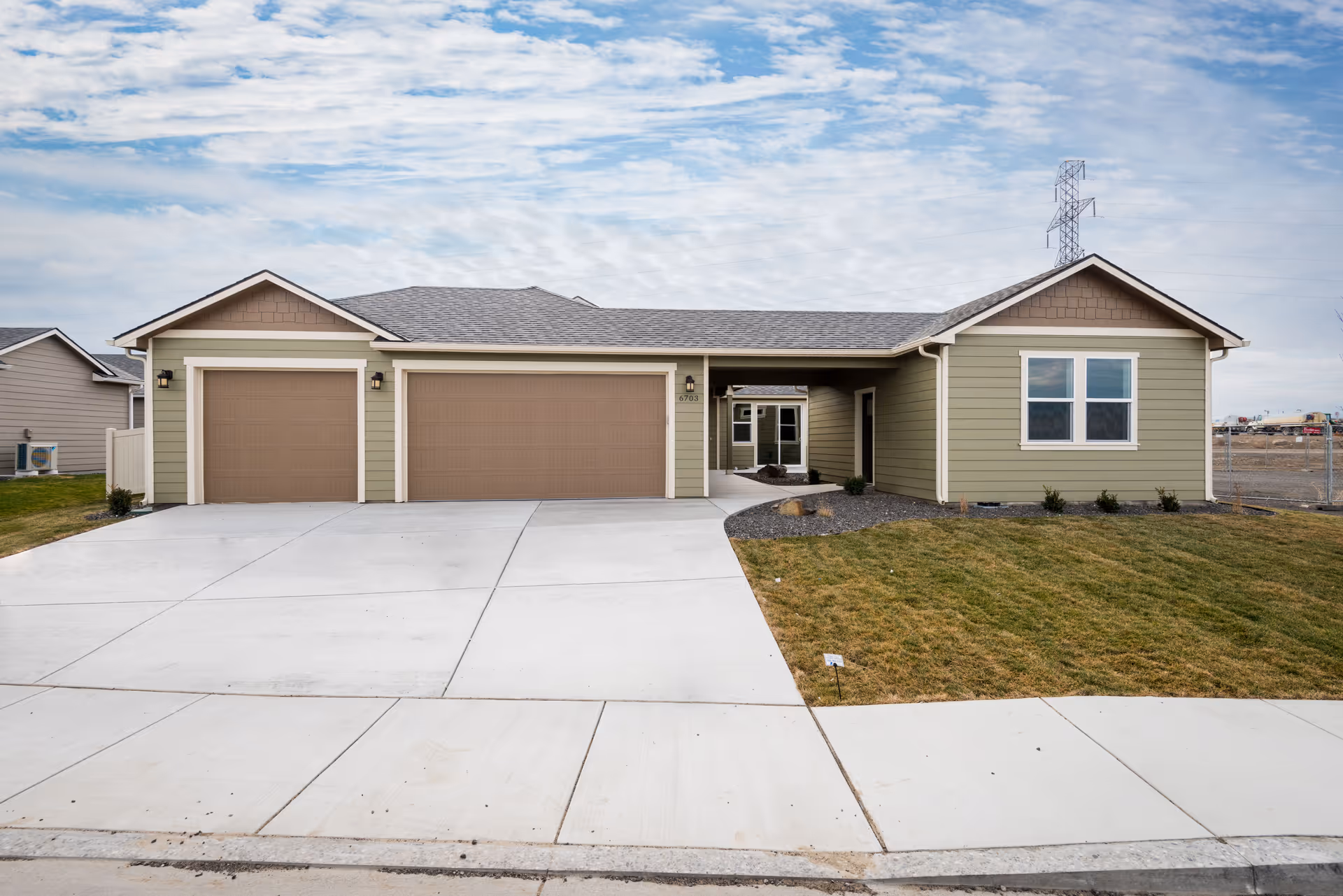 Front exterior view of a single-story residential building with a three-car garage, green siding, brown garage doors, and a concrete driveway. There is a small lawn area with some shrubs and a partly cloudy sky in the background.