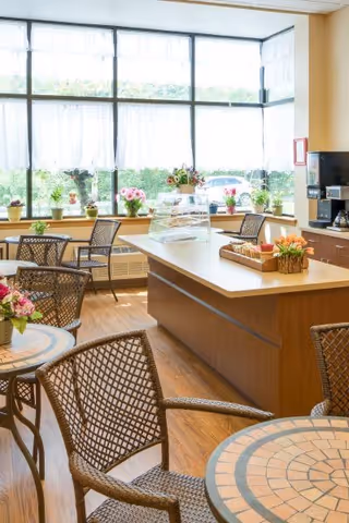 A bright senior dining area featuring round mosaic-top tables, wicker chairs, a central service island with flowers, and large windows with lace curtains.