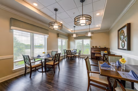 A bright dining room with several wooden tables and chairs, each table set with blue napkins and glassware. Large windows let in natural light and offer a view of greenery outside. The room features modern hanging light fixtures and a piano against the far wall with decorative items on top.