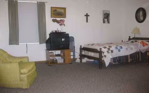 Senior living bedroom with bed, green chair, TV stand, and crucifix wall decor.