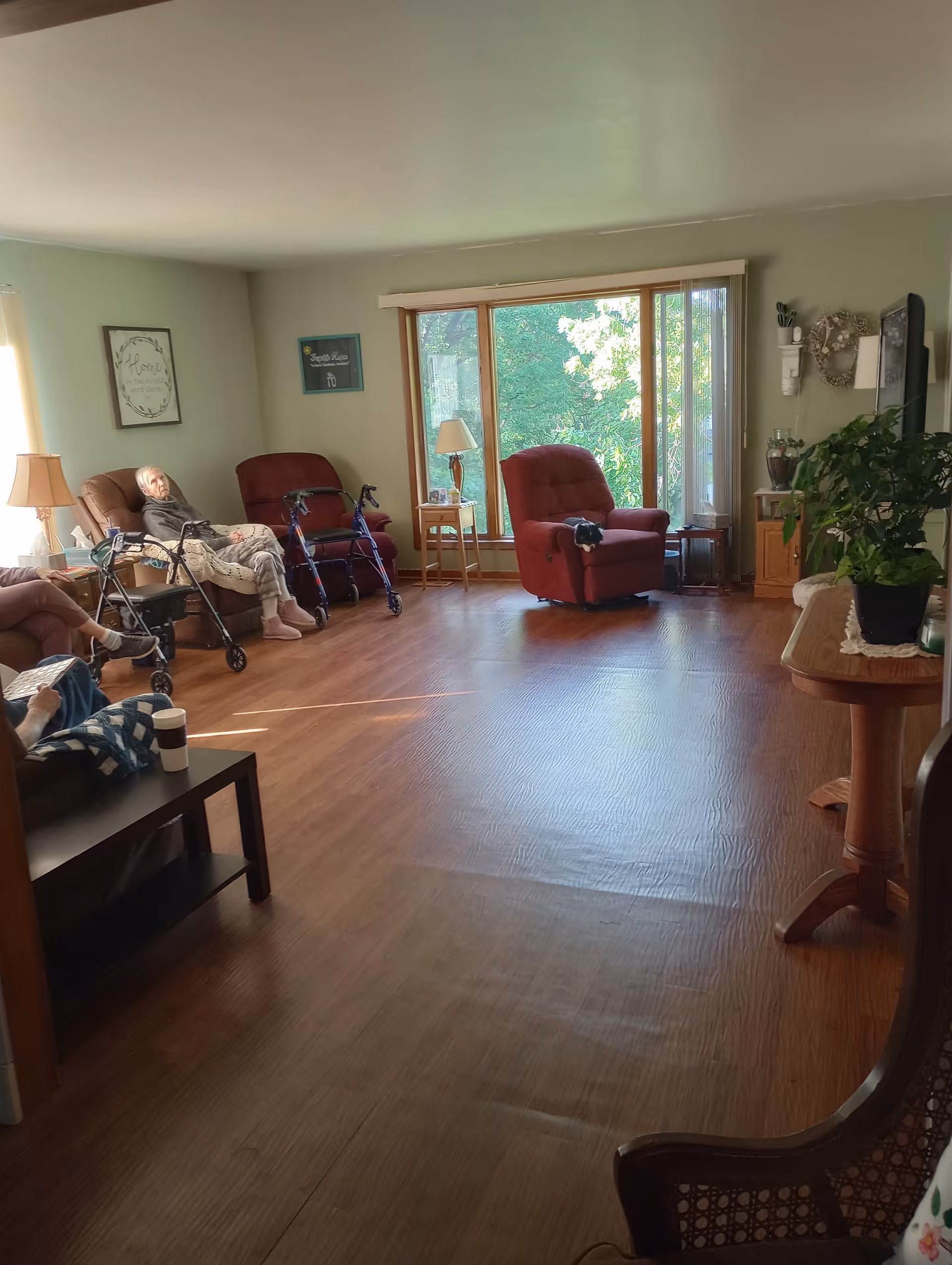A bright living room in an assisted living facility with wooden floors, large windows showing green trees outside, and several comfortable chairs including recliners. Two elderly individuals are seated in the chairs with walkers nearby. There is a small table with a lamp and a potted plant on a side table. The walls are light green with framed artwork.