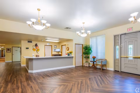 Interior view of a senior living facility lobby with a reception desk, decorative wall art, two chandeliers, a small seating area with two chairs and a table with flowers, large windows with blinds, and a double door exit.