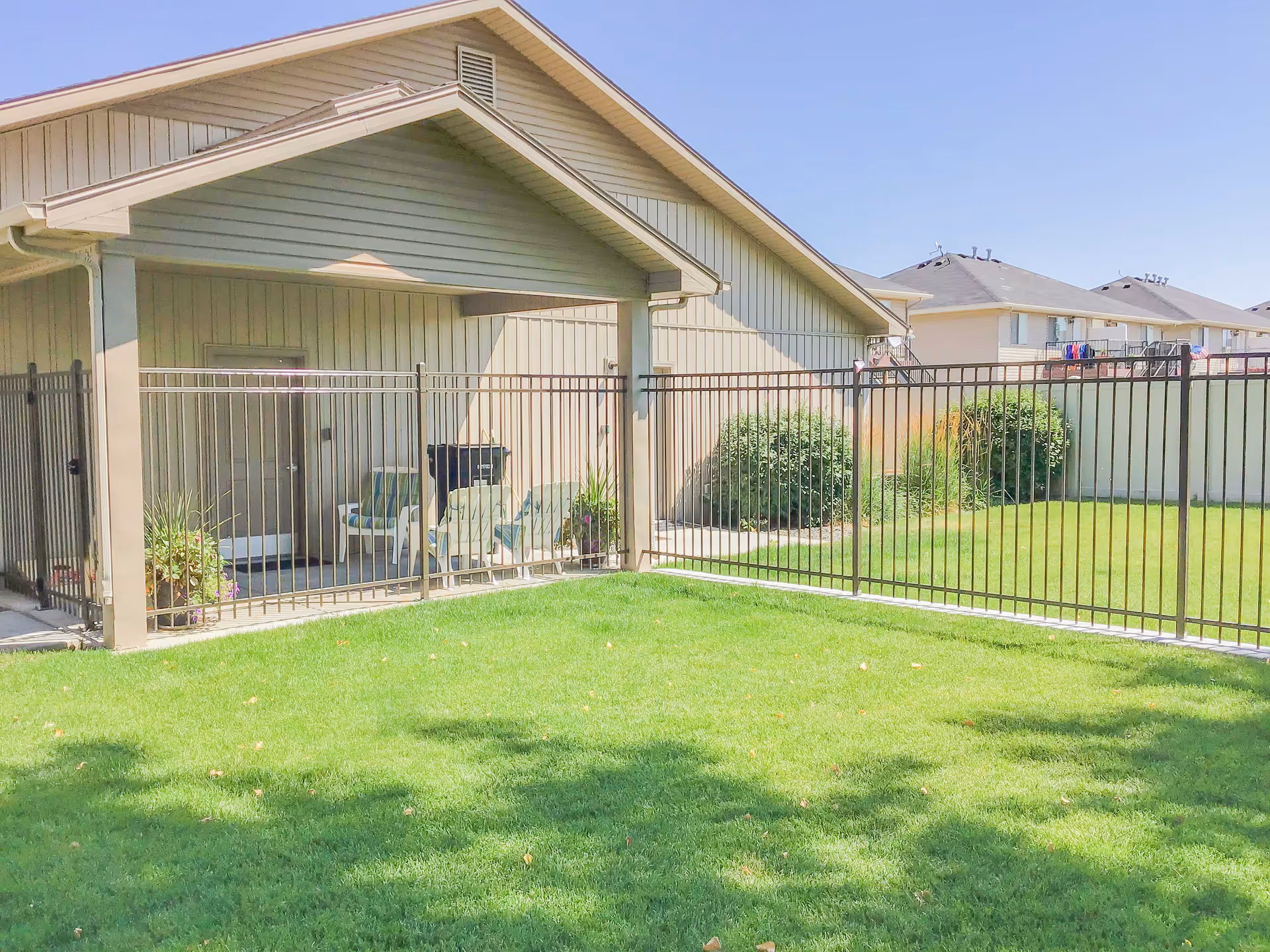Grassy fenced yard with a covered patio area, chairs, and potted plants next to a single-story building.