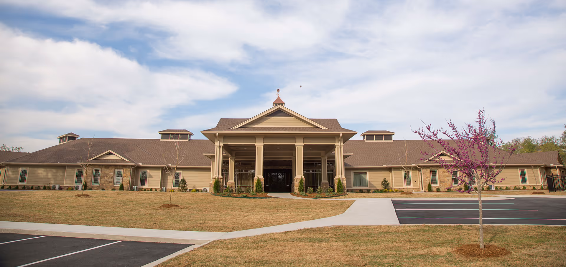 Front exterior of a single-story senior living facility with a central covered entrance, lawn, and parking area.