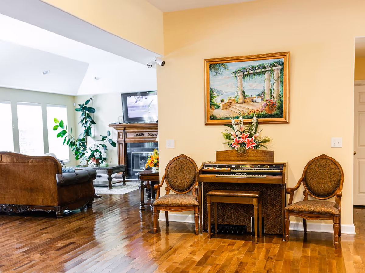 A cozy living room area with polished wooden floors, featuring a vintage organ with a floral arrangement on top, flanked by two ornate wooden chairs with upholstered seats. In the background, there is a brown leather sofa, a coffee table with a flower vase, a fireplace with a mounted TV above it, and large windows letting in natural light.