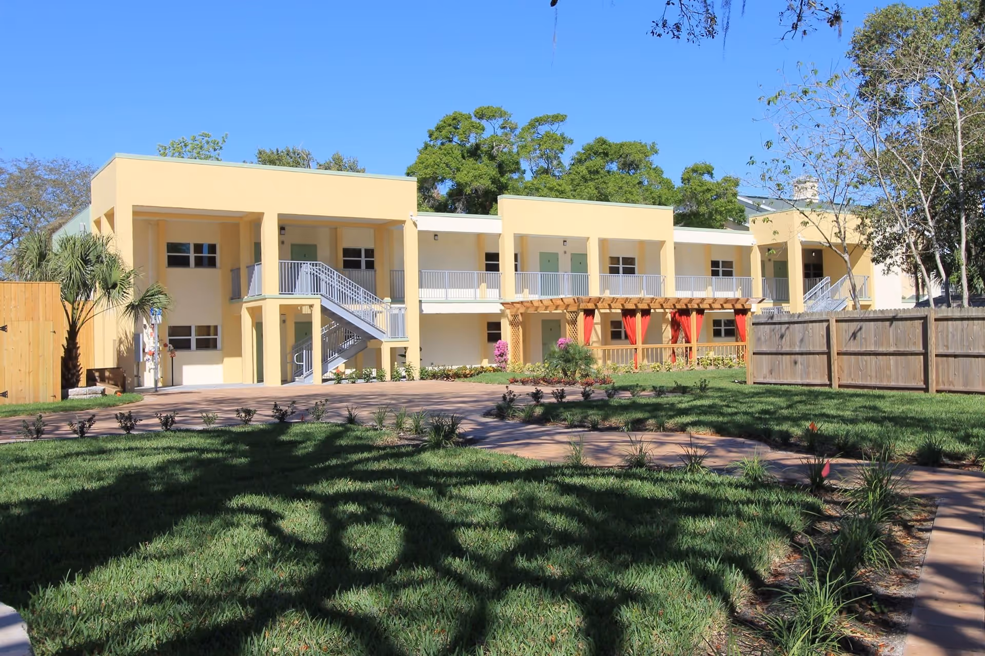 Exterior view of a two-story yellow senior living facility building with balconies, stairs, and a landscaped garden with green grass and plants under a clear blue sky.