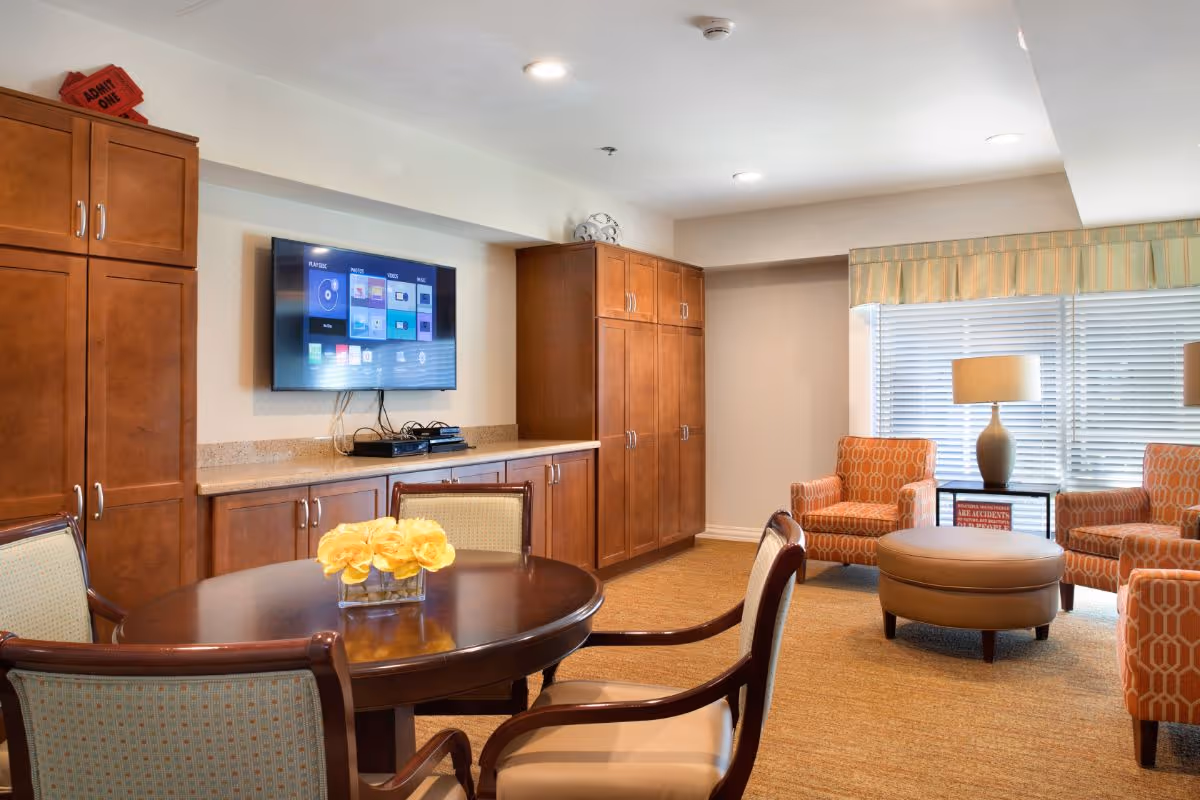 A cozy common area with a round wooden table and four chairs, a flat-screen TV mounted on the wall above wooden cabinets, and a seating area with two orange patterned armchairs, a round ottoman, and a table lamp near a window with blinds and a striped valance.