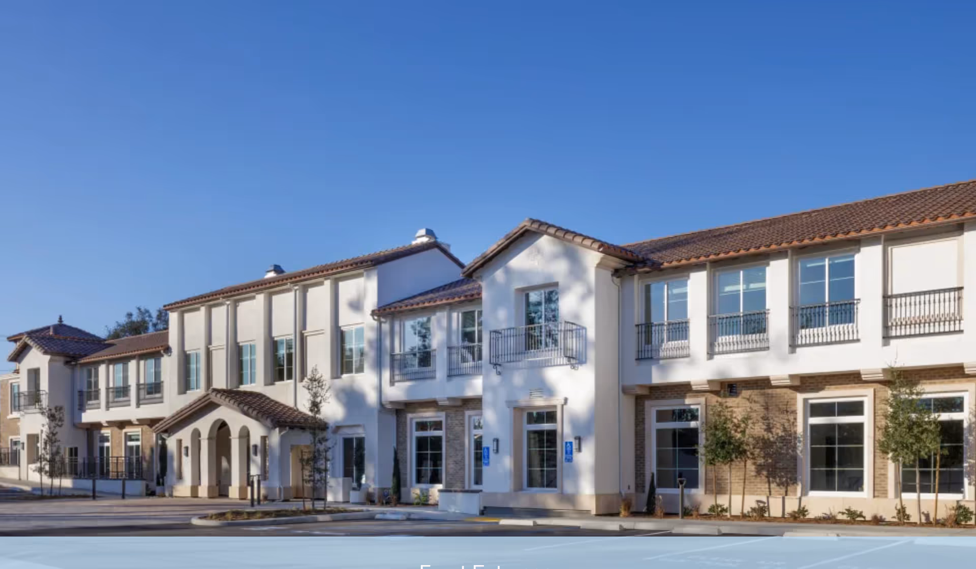 Front exterior of a two-story Mediterranean-style senior living building with balconies, arched entrance, and clear blue sky.