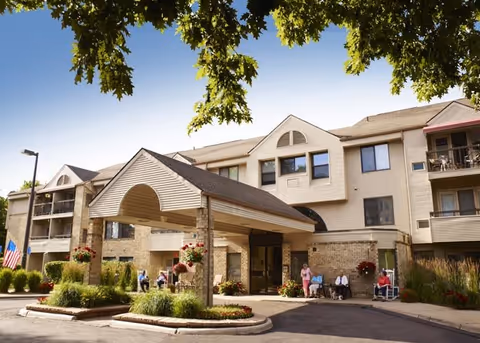Exterior view of a senior living facility named American House Carpenter with a covered entrance, landscaped greenery, and several people sitting outside near the entrance under a clear blue sky.