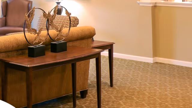 A cozy interior space featuring a wooden console table with two decorative metal sculptures on top, positioned behind a cushioned sofa. The room has beige walls and patterned carpet flooring.