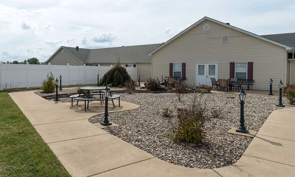 Outdoor courtyard with paved walkways, lamp posts, picnic tables and patio seating in front of a beige single-story senior living building.