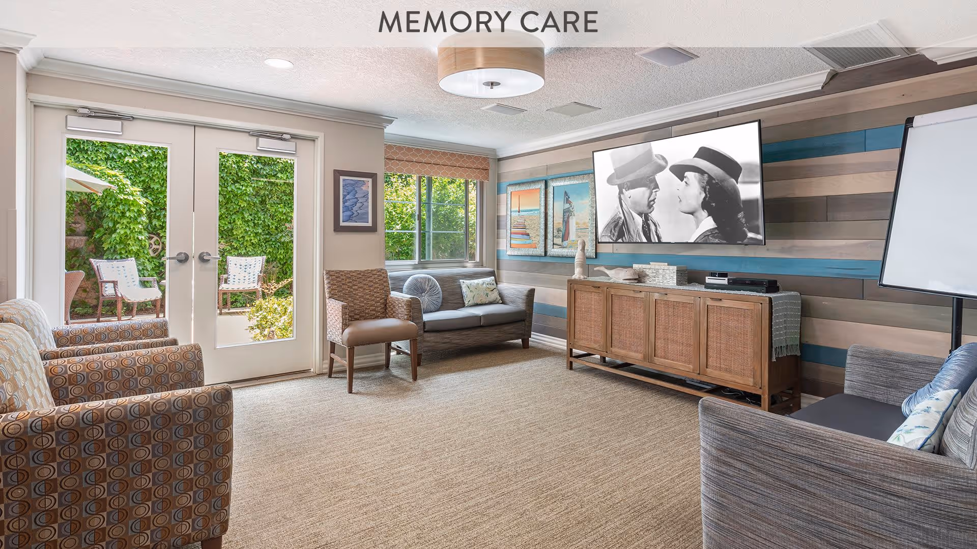 A bright and cozy memory care living room with patterned armchairs, a sofa, and a chair arranged around a large flat-screen TV mounted on a striped wooden accent wall. There is a cabinet beneath the TV, a window letting in natural light, and double glass doors leading to an outdoor seating area with greenery.