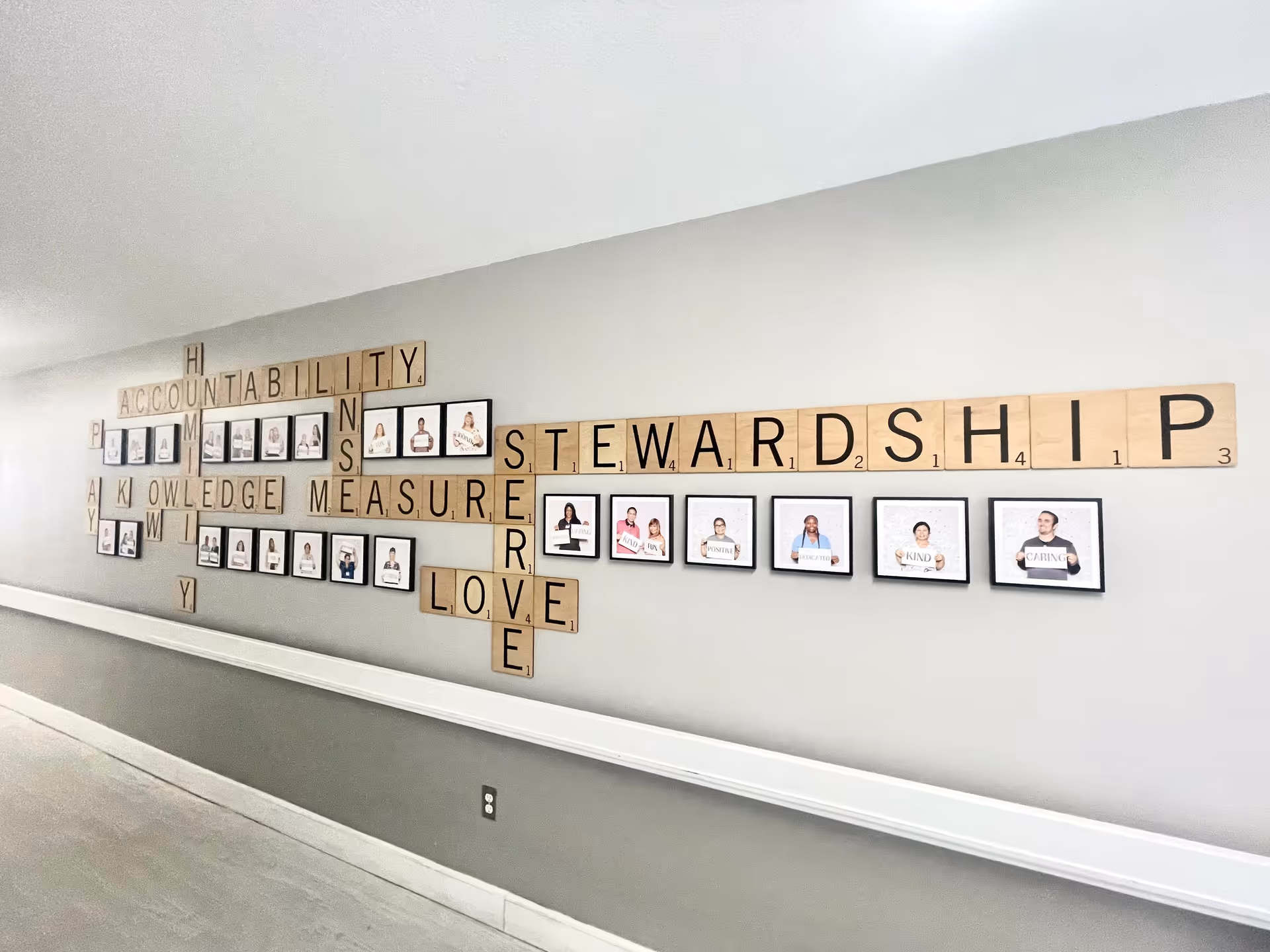 A hallway wall decorated with large Scrabble-style letter tiles spelling words like ACCOUNTABILITY, STEWARDSHIP, KNOWLEDGE, MEASURE, LOVE, and SERVE. Below and between the words are framed photos of people holding signs with positive words such as KIND and CARING.
