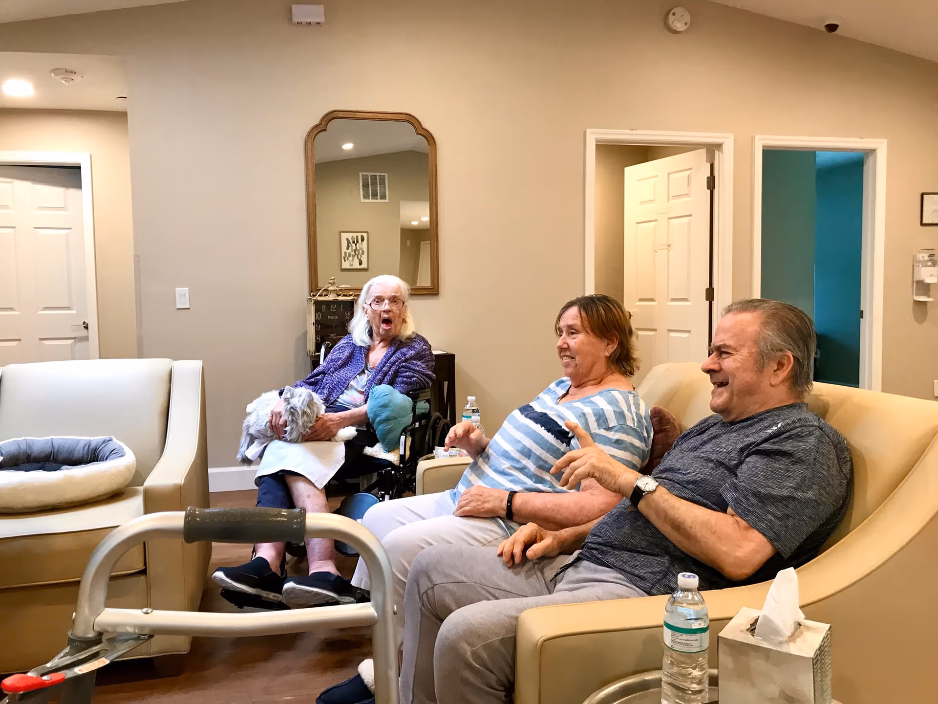 Three seniors sitting and laughing in a comfortable common living room with chairs, a walker, and a water bottle.