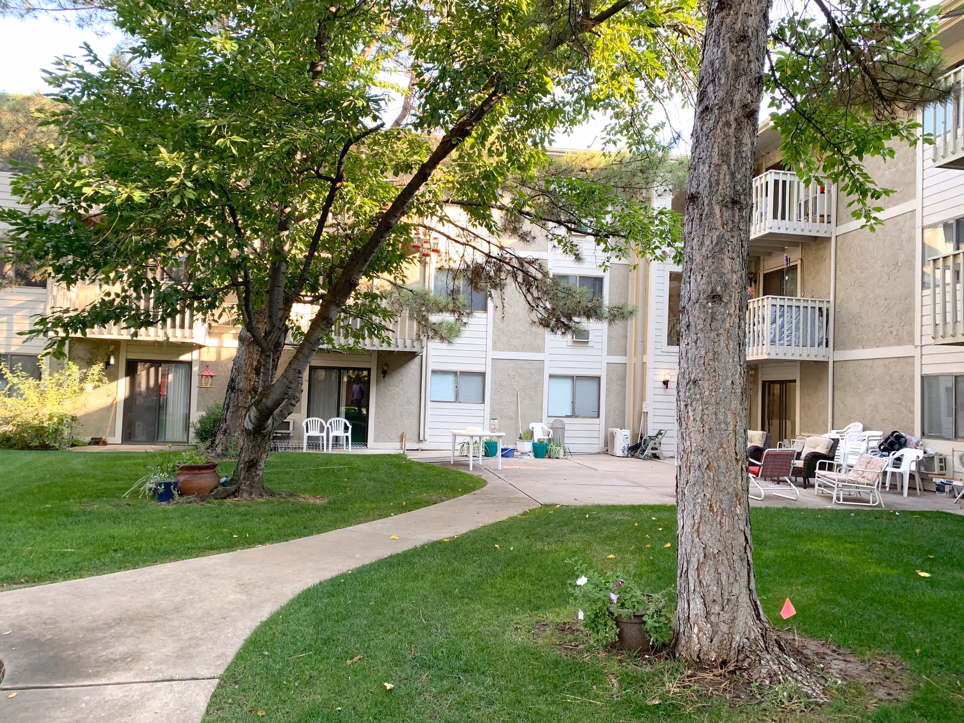 Outdoor courtyard area of a retirement community with green grass, trees, and a paved walkway. The building surrounding the courtyard has multiple balconies and sliding glass doors. There are several white and brown chairs arranged on the patio area.