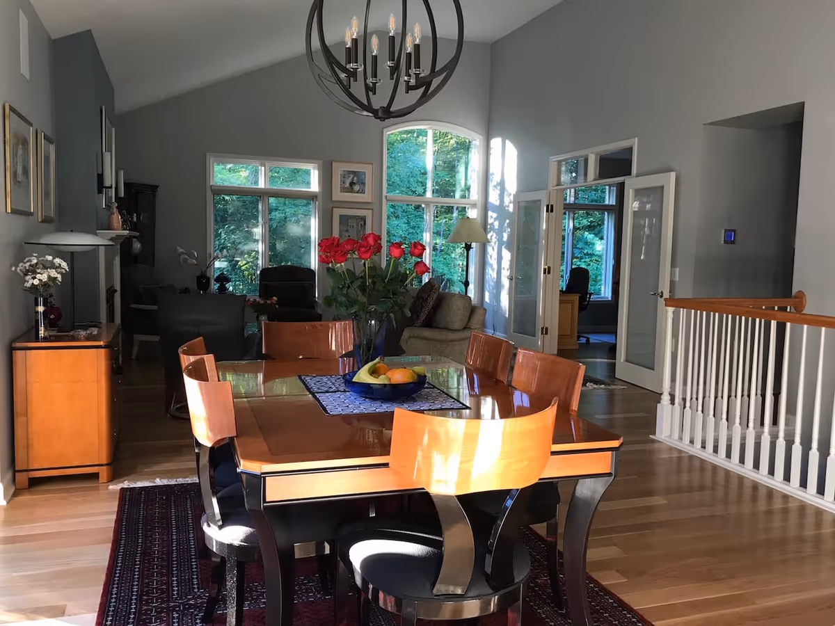 Sunlit open dining area with a wooden dining table and chairs, a vase of red roses, hardwood floors and adjacent living room with large windows.