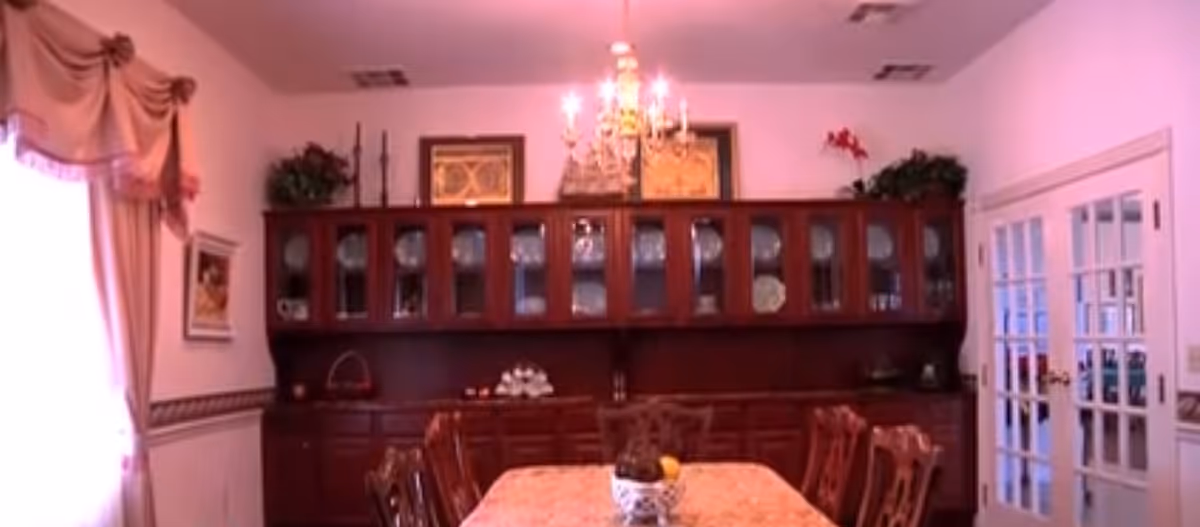 Dining room with a wooden table and chairs, a chandelier hanging from the ceiling, a large wooden cabinet with glass doors displaying dishes, and a window with draped curtains on the left side.