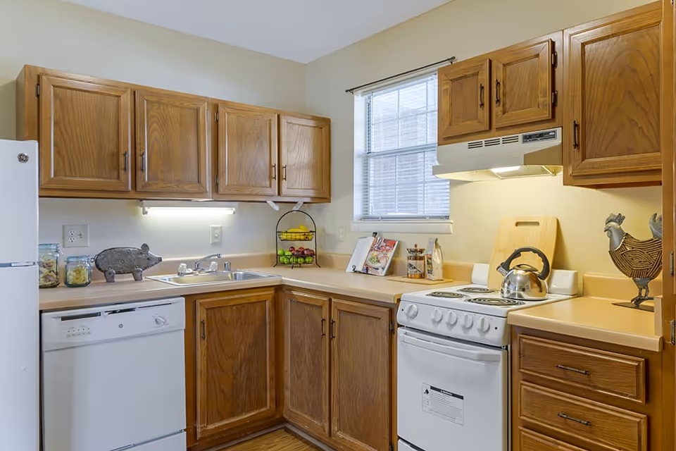 A kitchen with wooden cabinets, a white refrigerator, a white dishwasher, a white stove with a kettle on it, a sink, and a window with blinds. The countertop has a cutting board, a cookbook, a fruit basket, and decorative items including a pig and a rooster.