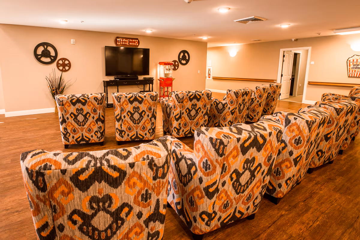 A cozy media room with multiple patterned armchairs arranged in rows facing a large flat-screen TV mounted on the wall. The room features wood flooring, beige walls, decorative film reels on the wall, a popcorn machine, and a lighted sign above the TV that reads 'CINEMA'.