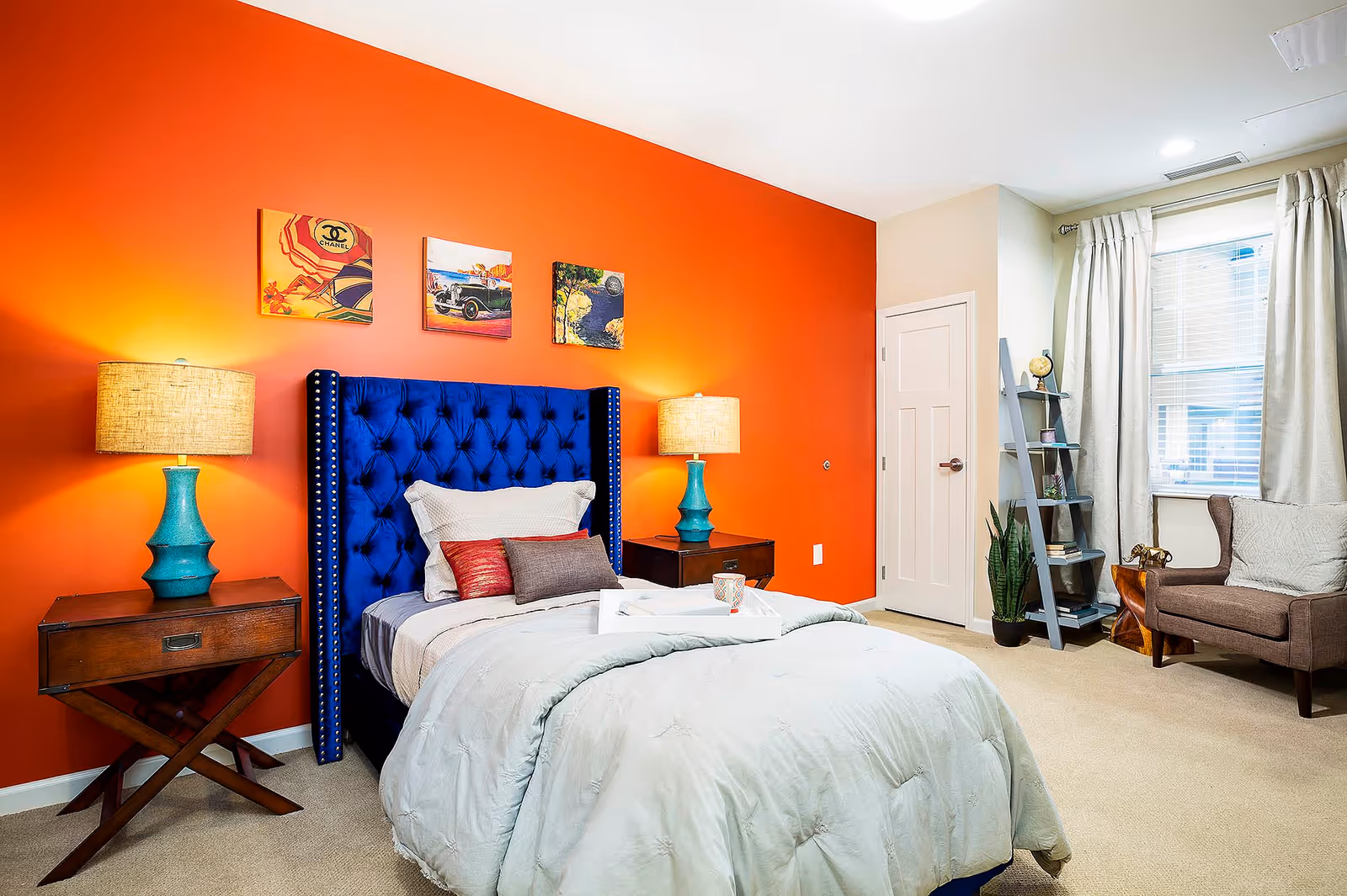 A bedroom with a vibrant orange accent wall behind a blue tufted headboard bed. The bed is made with light-colored bedding and several pillows. On each side of the bed are wooden nightstands with turquoise lamps. Above the bed are three colorful framed artworks. To the right, there is a window with beige curtains, a gray ladder-style bookshelf with decorative items, a potted plant, and a brown upholstered armchair with a cushion.