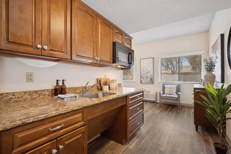 A kitchen area with wooden cabinets, granite countertops, a sink, and a microwave. The kitchen opens into a small sitting area with a gray armchair, a wooden dresser, two windows with blinds, and decorative plants and artwork on the walls.