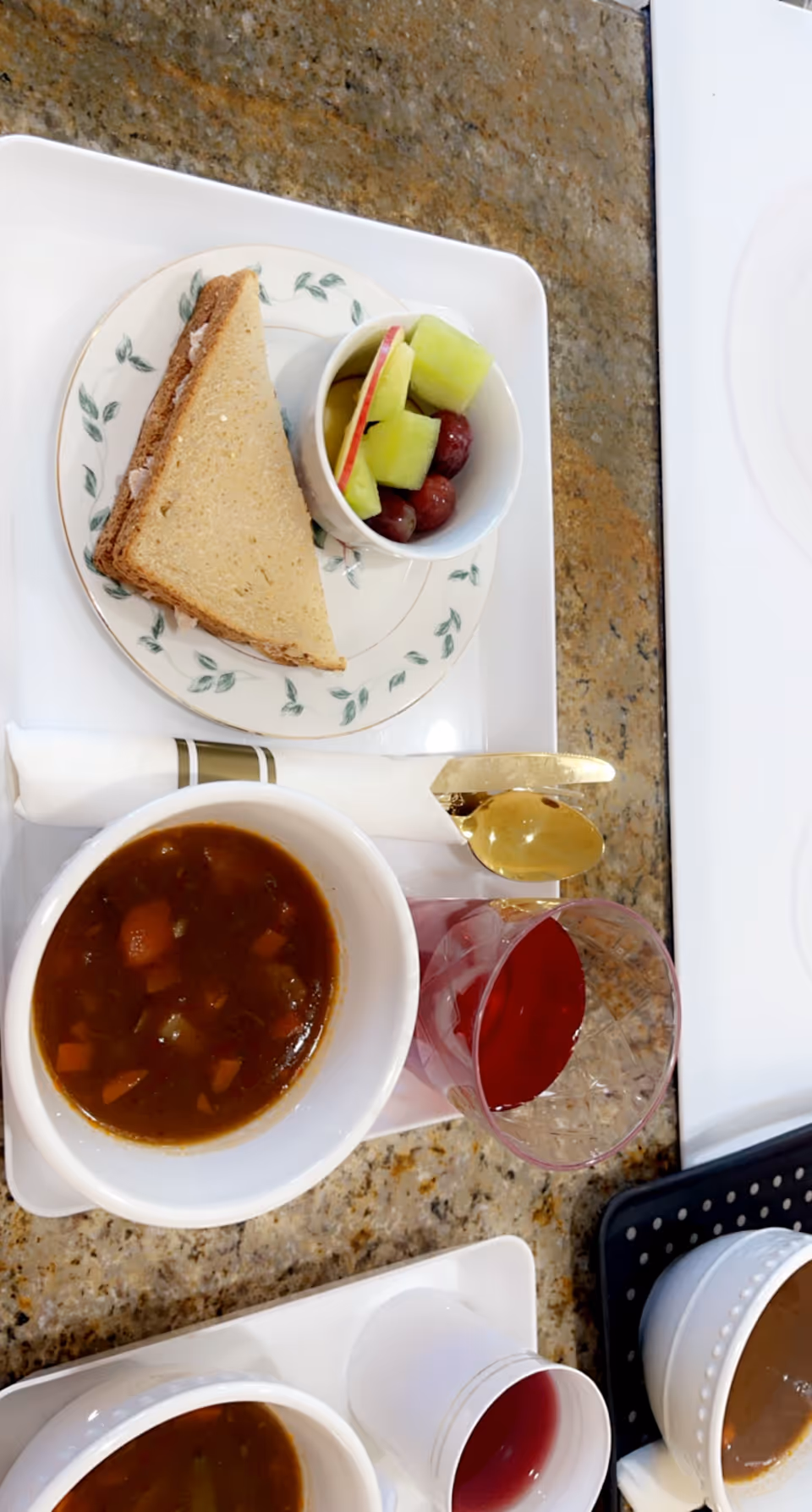A meal tray on a granite countertop with a bowl of vegetable soup, a sandwich on a floral plate, a small cup of mixed fruit including grapes, melon, and apple slices, a glass of red juice, and a wrapped napkin with a gold spoon.