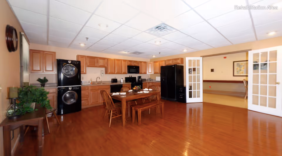 A spacious kitchen area with wooden cabinets, black appliances including a refrigerator, microwave, stove, and a stacked washer and dryer. A wooden dining table with chairs and a bench is set in the center of the room. The floor is wooden, and there are double glass doors leading to another room. A small table with plants is on the left side near a window.