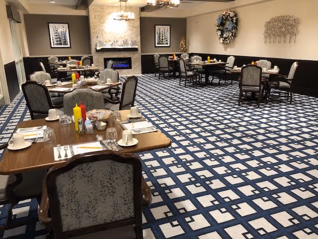 A dining room with multiple tables set for meals, each table having chairs, cups, plates, utensils, and condiments like ketchup and mustard. The room features a patterned carpet, a stone fireplace with a modern electric fire, framed artwork on the walls, and decorative wreaths and wall art.