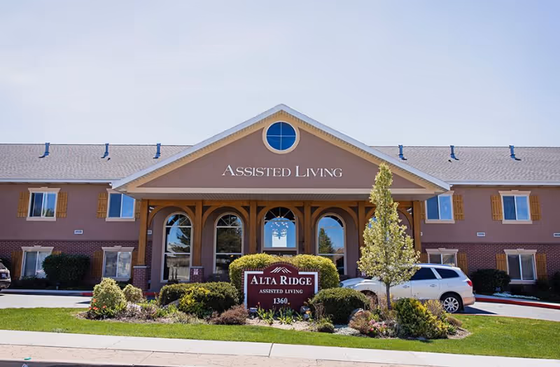 Front exterior view of Alta Ridge Assisted Living of Holladay building with a large covered entrance, windows, and landscaping including bushes and a small tree. A sign in front reads 'Alta Ridge Assisted Living 1360'. A white vehicle is parked to the right side of the building.