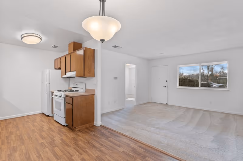 Interior view of a senior living facility showing a small kitchen area with wooden cabinets, a white refrigerator, and a white stove. Adjacent to the kitchen is a carpeted living room area with a window and a door. The walls are white, and the floor in the kitchen area is wood laminate. A hanging light fixture is visible above the kitchen space.