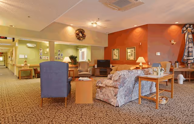 A cozy living room area in a senior living facility with patterned carpet, floral and solid upholstered sofas and chairs, wooden side tables with lamps, framed artwork on the walls, and a television on a stand. The walls are painted in warm tones of green and orange, and there is a hallway visible to the left.