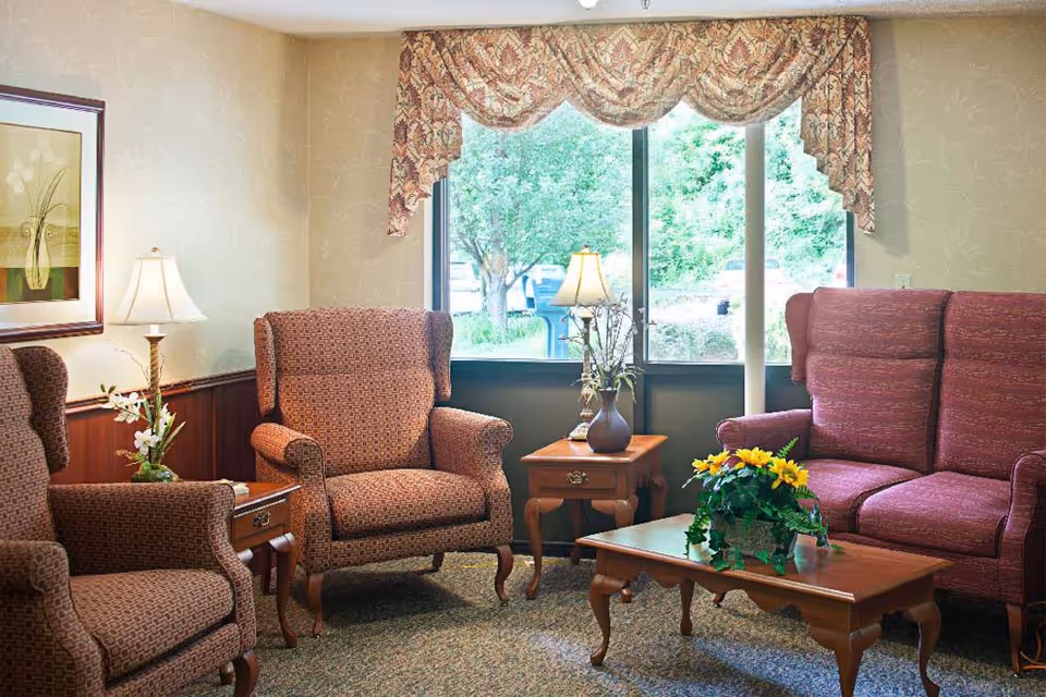 A cozy living room area with three upholstered armchairs and a loveseat in a patterned fabric. There are two wooden side tables with lamps and decorative plants, and a wooden coffee table with a potted plant featuring yellow flowers. A large window with floral valances lets in natural light and shows greenery outside. The walls have a light patterned wallpaper and a framed floral artwork.