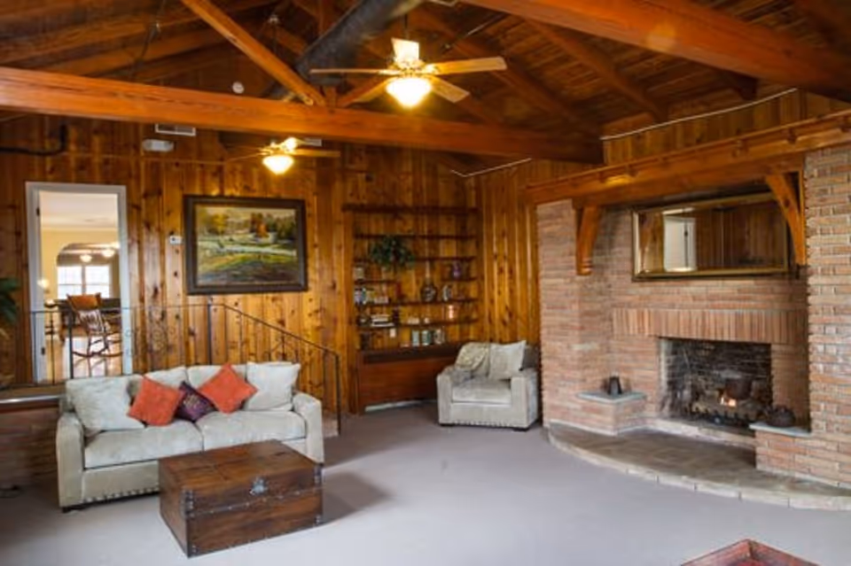 A cozy living room with wood-paneled walls and exposed wooden beams on the ceiling. The room features a beige sofa with red and purple cushions, a matching armchair, a wooden chest used as a coffee table, and a brick fireplace with a mirror above it. There is a built-in wooden bookshelf with decorative items and a painting on the wall. A doorway leads to another room with a dining area visible.