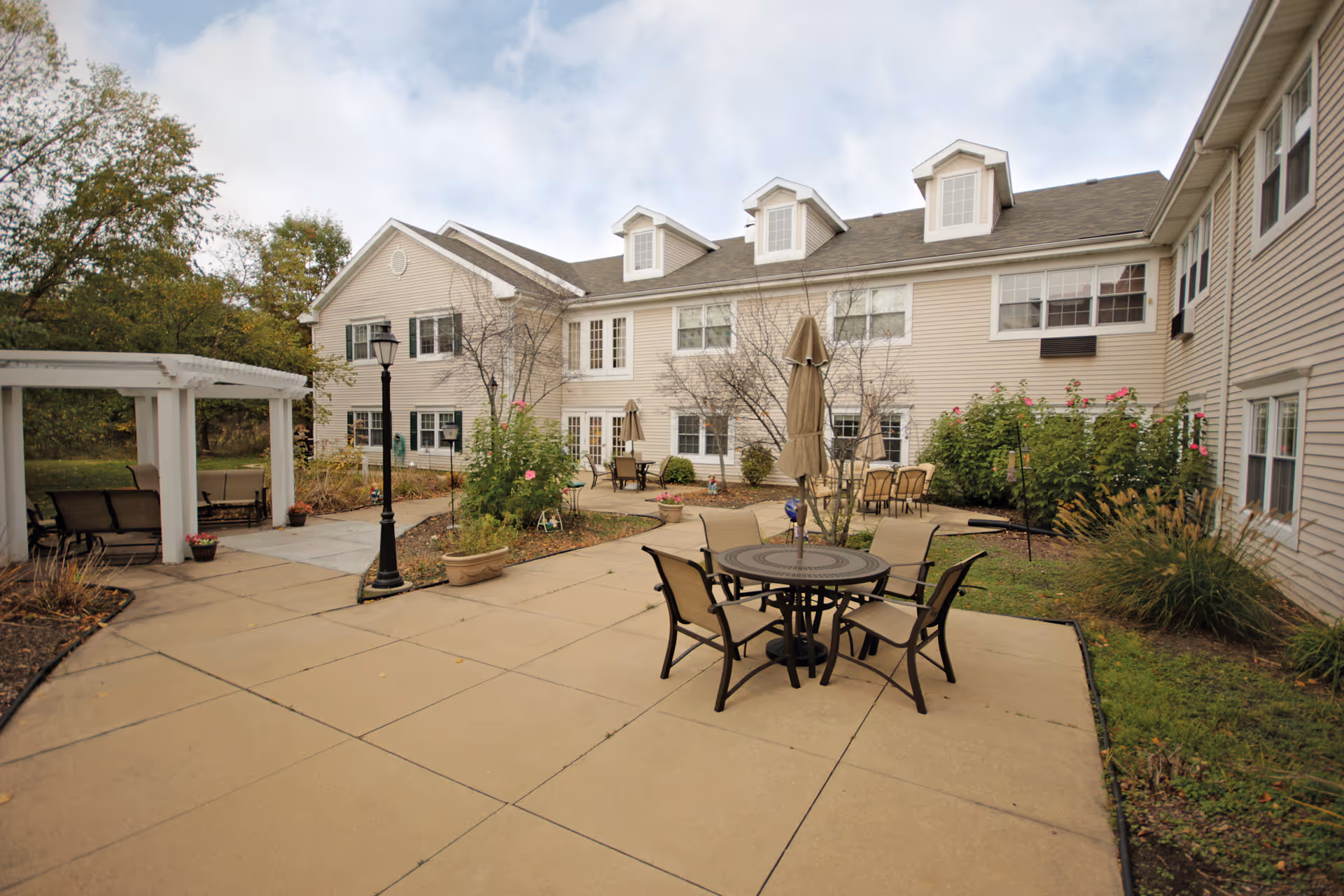 Outdoor courtyard patio at an assisted living facility with round tables, umbrellas, a pergola and a beige two-story building in the background.