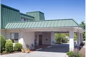 Exterior view of a building with a green metal roof and a covered entrance supported by stone pillars. There are shrubs and small plants around the entrance area under a clear blue sky.