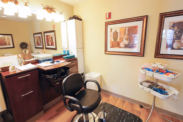 Interior view of a salon area in a senior living facility featuring a black salon chair in front of a counter with a sink, cabinets, and various hair care products. The room has wooden flooring, beige walls with two framed pictures, and a rolling cart with hair accessories and tools.
