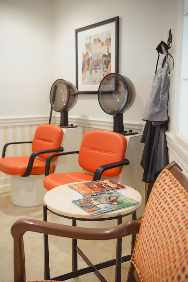Small salon area with two hooded hair dryers, orange chairs, a round table with magazines, and aprons hanging on the wall.