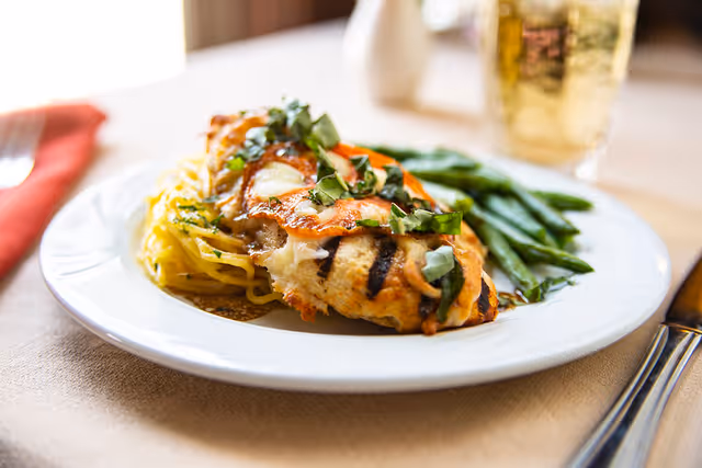 A plate of grilled chicken topped with melted cheese and fresh basil, served with green beans and a side of spaghetti squash, with a glass of iced tea in the background.