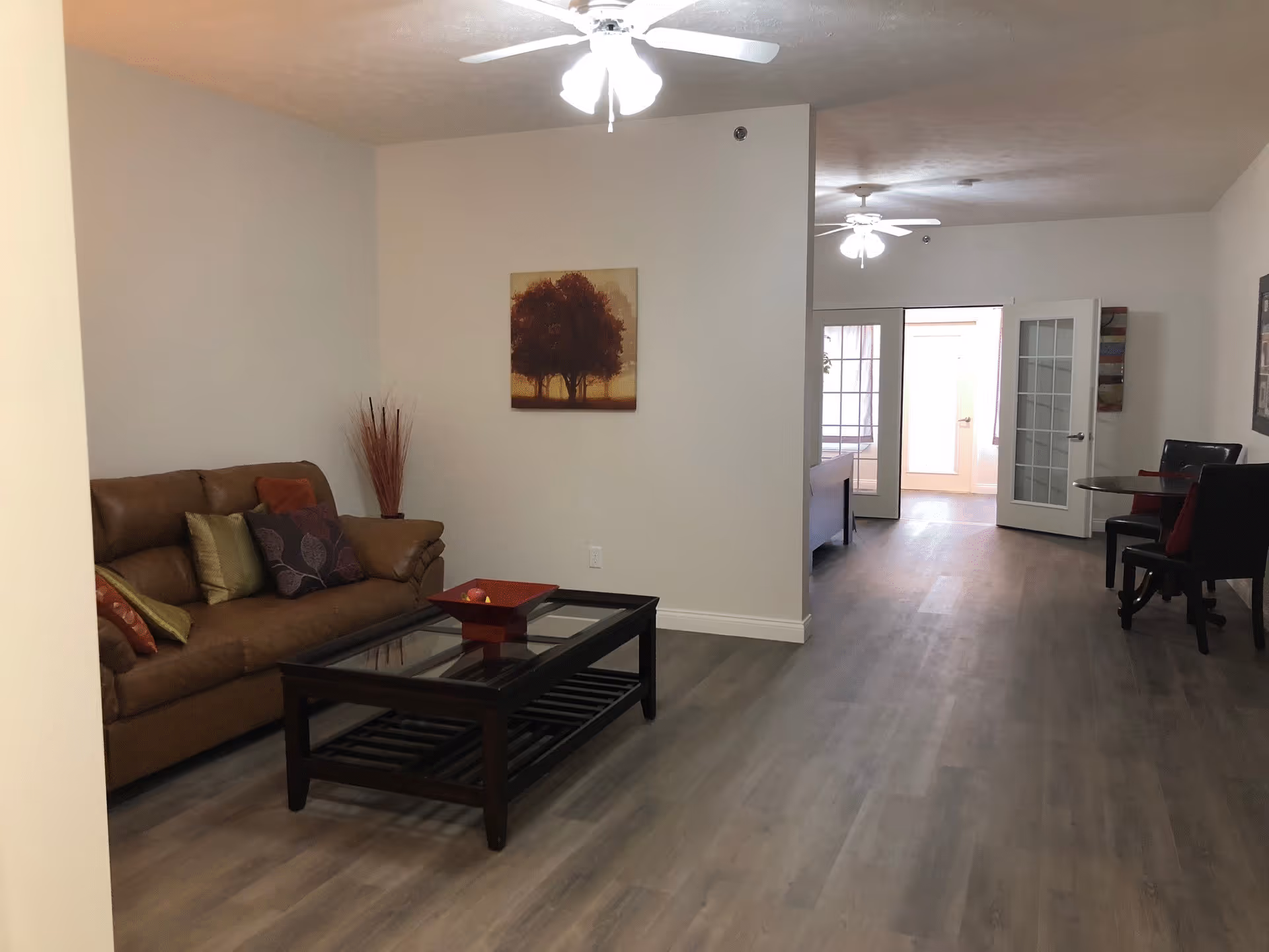 Interior view of a living area in an assisted living facility featuring a brown leather sofa with multiple cushions, a glass-top coffee table with a decorative tray, a wall painting of trees, and ceiling fans with lights. In the background, there is a small dining area with a round table and chairs, and a hallway leading to other rooms with glass-paneled doors.